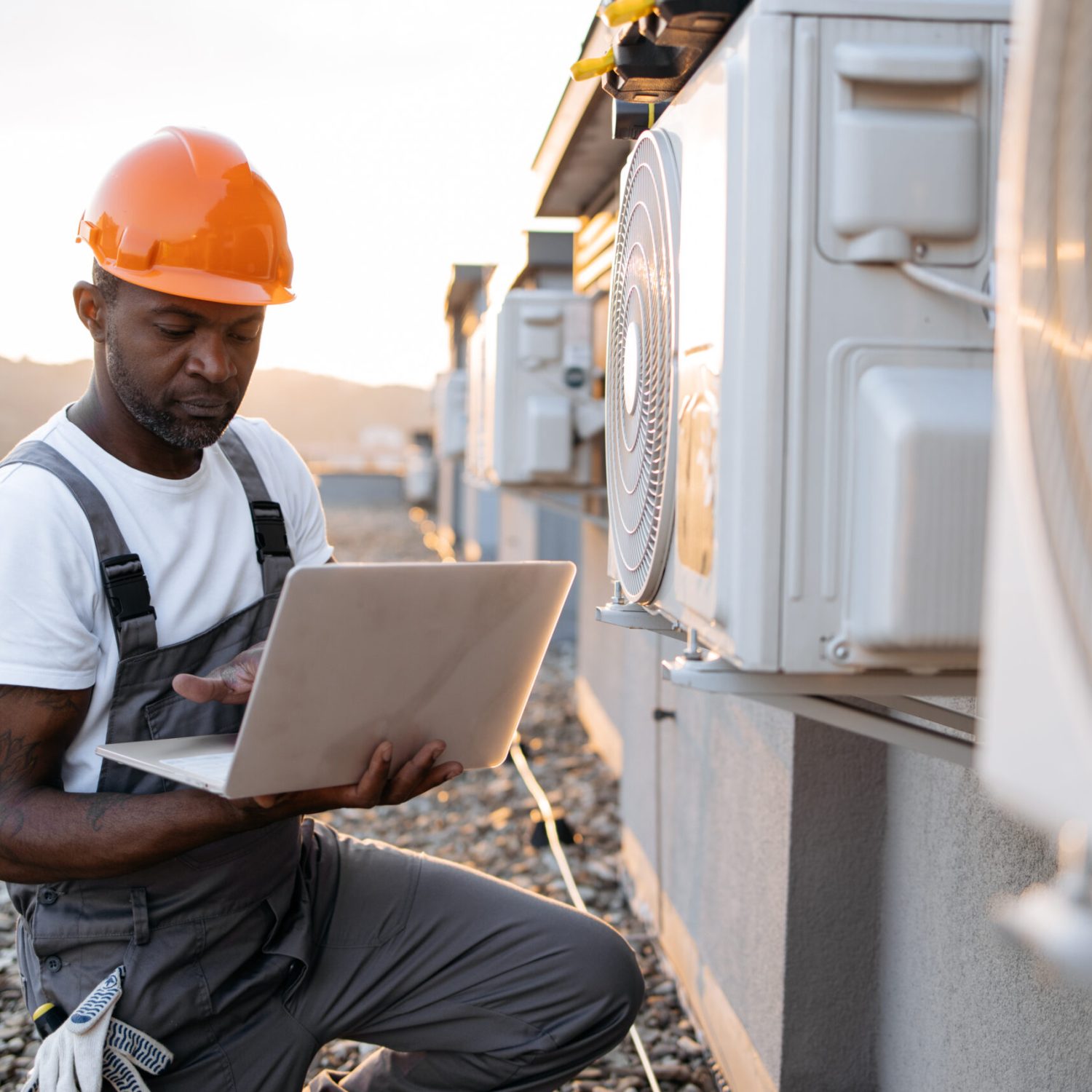 Concentrated male technician using wireless laptop while kneeling near air conditioner on rooftop. African american specialist typing information on modern gadget about AC unit in work report.